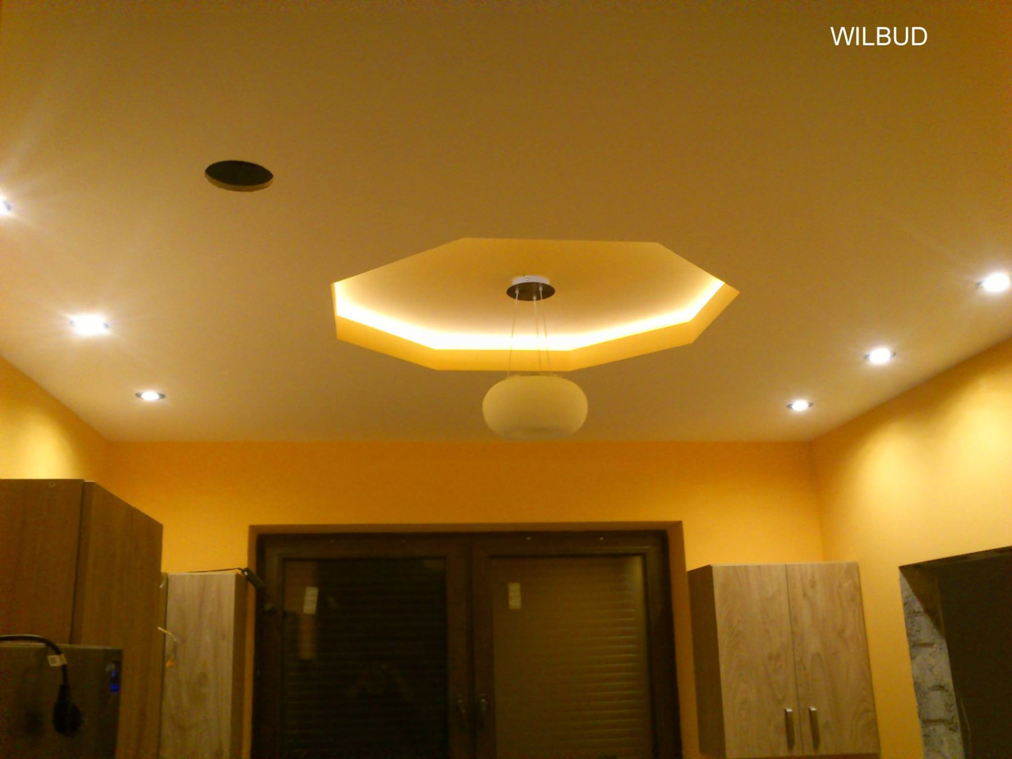 Żółta kuchnia z podwieszanym sufitem w kształcie ośmiokąta z oświetleniem LED i okrągłą lampą wiszącą, widoczne również halogeny i meble.