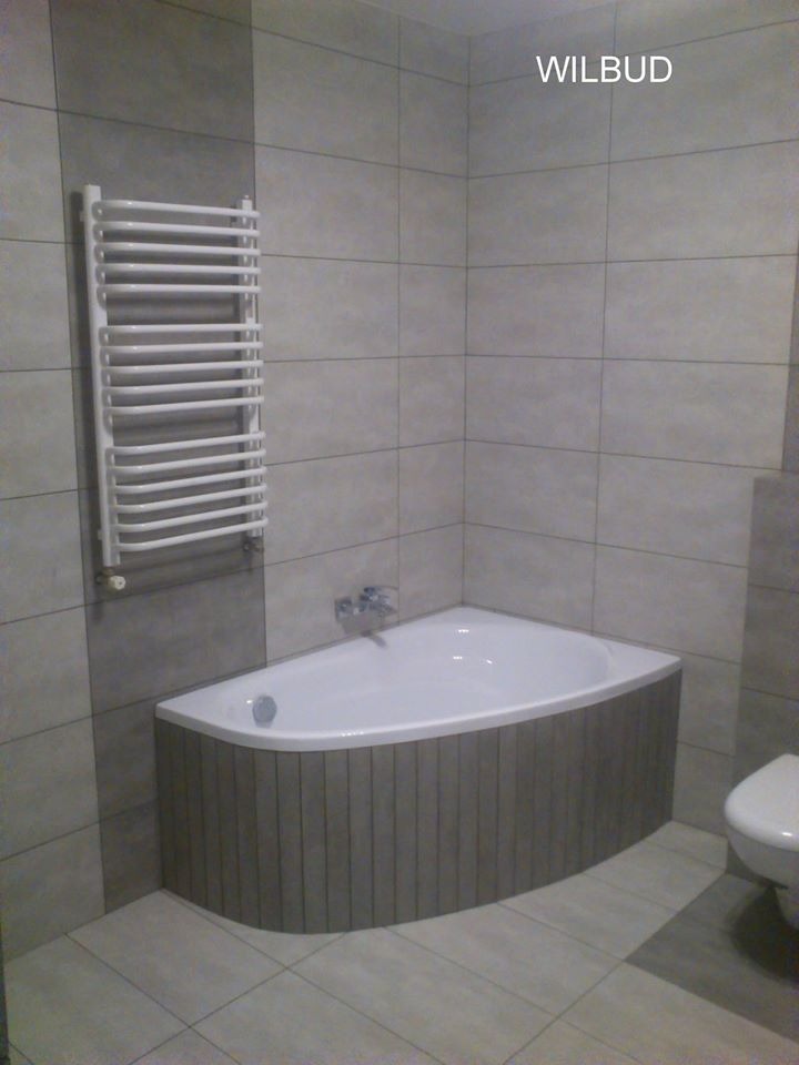 Wykończona łazienka z narożną wanną obudowaną pionowymi panelami, grzejnikiem drabinkowym na ścianie i płytkami w odcieniach szarości, widoczny fragment toalety.