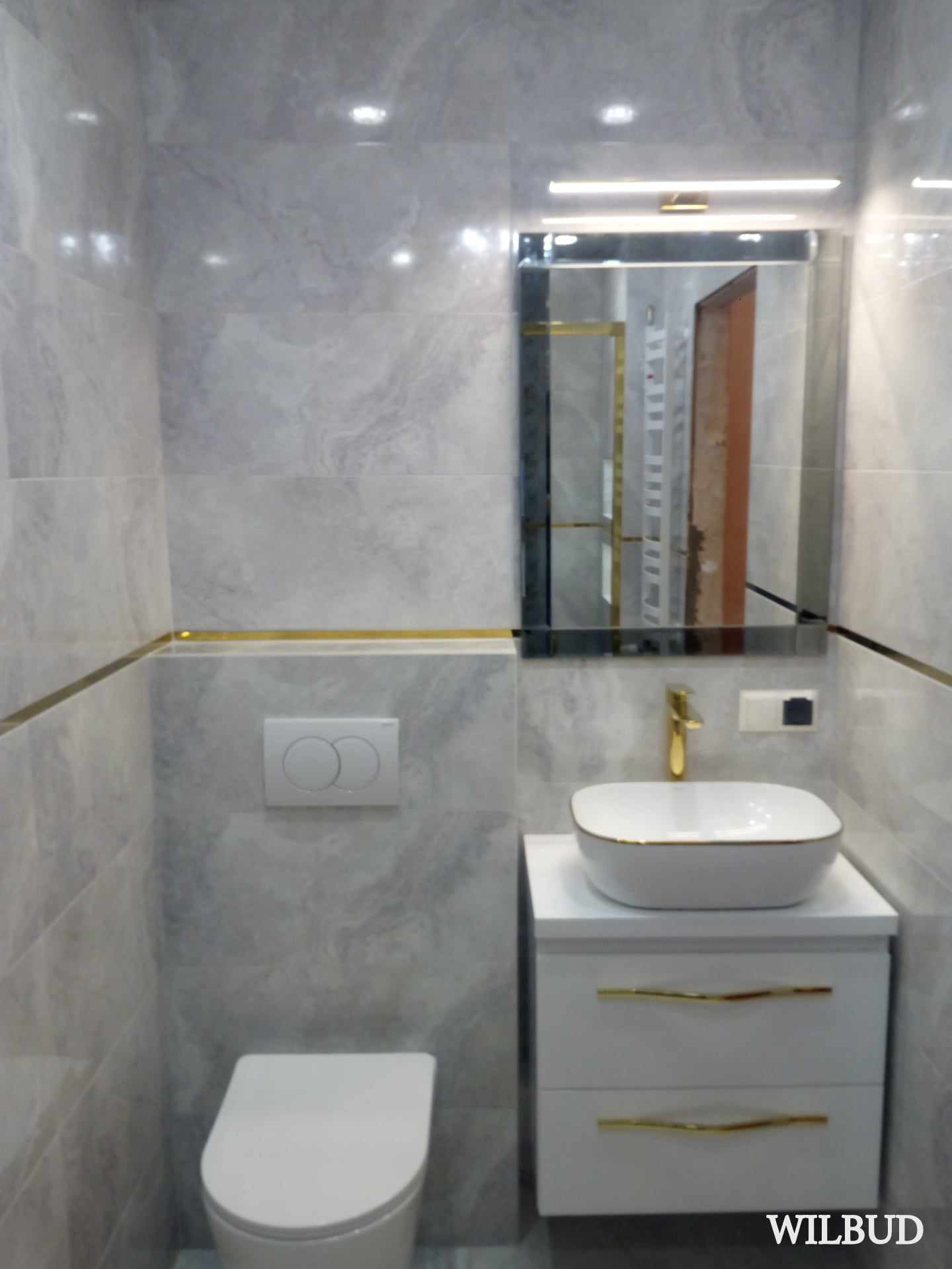 Wykończona łazienka z marmurowymi płytkami, złotymi akcentami, umywalką nablatową na białej szafce z dwiema szufladami i lustrem z podświetleniem LED.