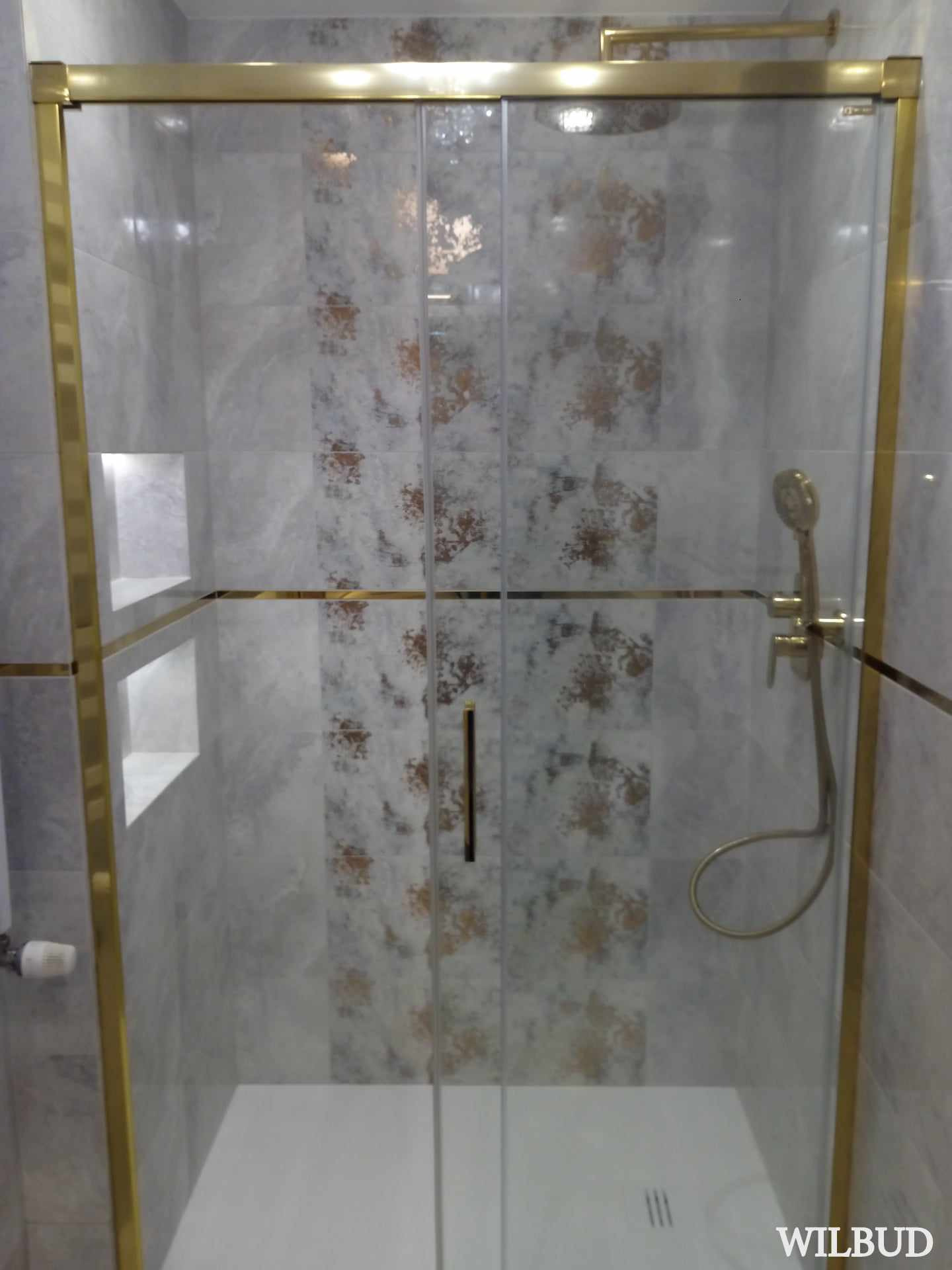 Szklana kabina prysznicowa z przesuwnymi drzwiami, wykończona złotymi profilami, zamontowana w łazience z szarymi płytkami o wzorzystej fakturze. W tle widoczna wnęka w ścianie.