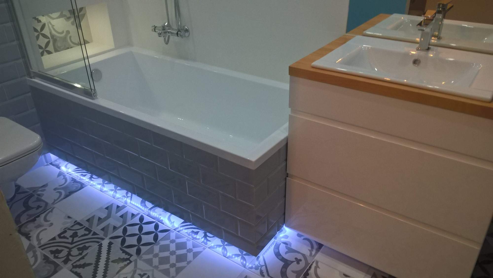 Nowoczesna łazienka z wanną obudowaną szarymi płytkami typu metro, podświetleniem LED pod wanną, wzorzystą podłogą i białą szafką z umywalką na drewnianym blacie.