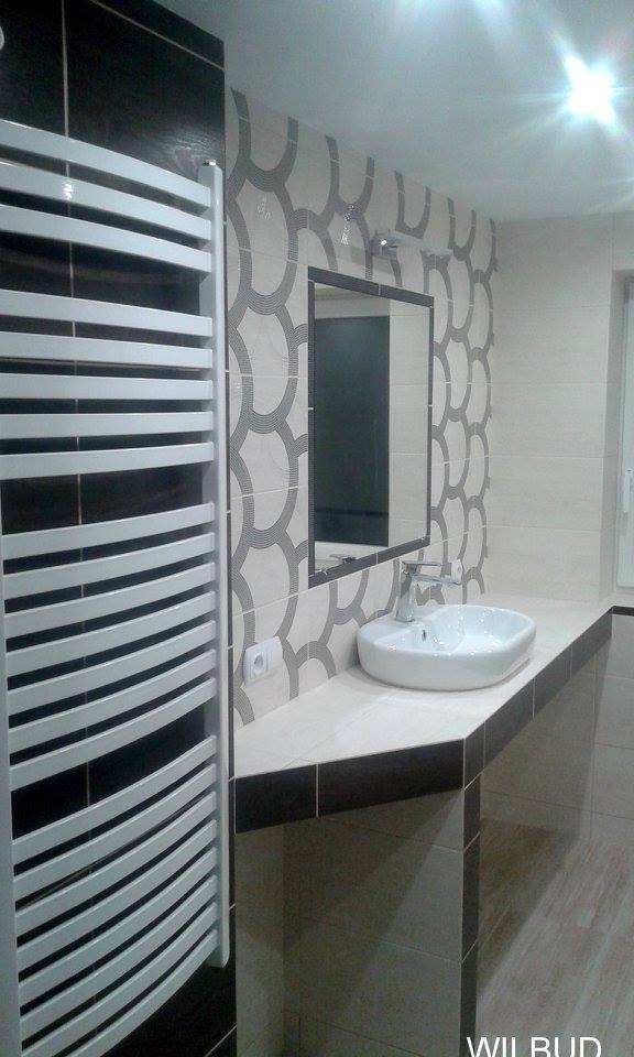 Wykończona łazienka z umywalką nablatową, grzejnikiem drabinkowym i wzorzystymi płytkami w odcieniach szarości i bieli, widoczny fragment lustra i gniazdko elektryczne.