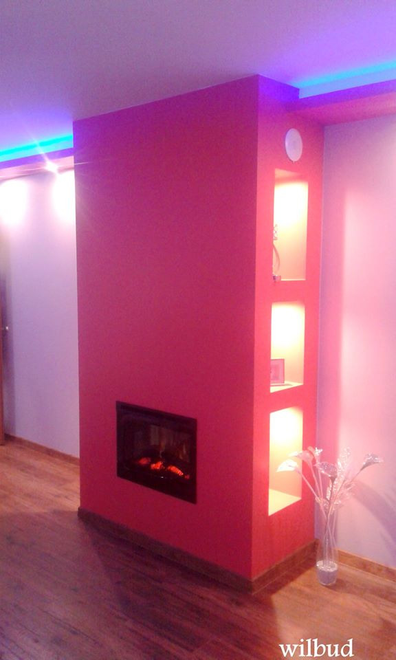 Czerwona ściana działowa z elektrycznym kominkiem i trzema podświetlanymi wnękami, oświetlenie LED na suficie, drewniana podłoga.