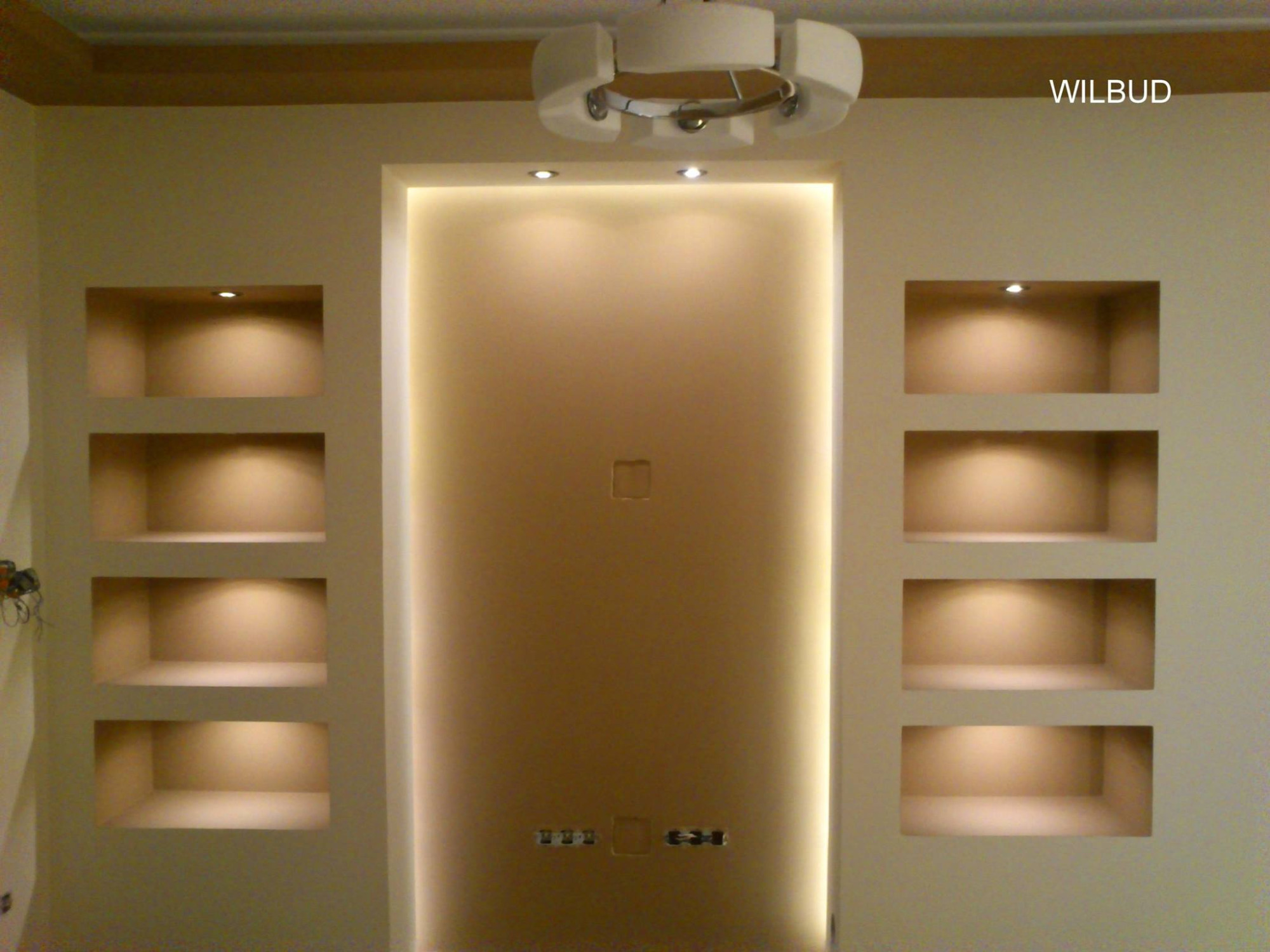 Ściana z wnękami oświetlonymi punktowo, centralna część ściany z podświetleniem LED po bokach, górne oświetlenie sufitowe, kremowa kolorystyka ścian, widoczny fragment instalacji elektrycznej.