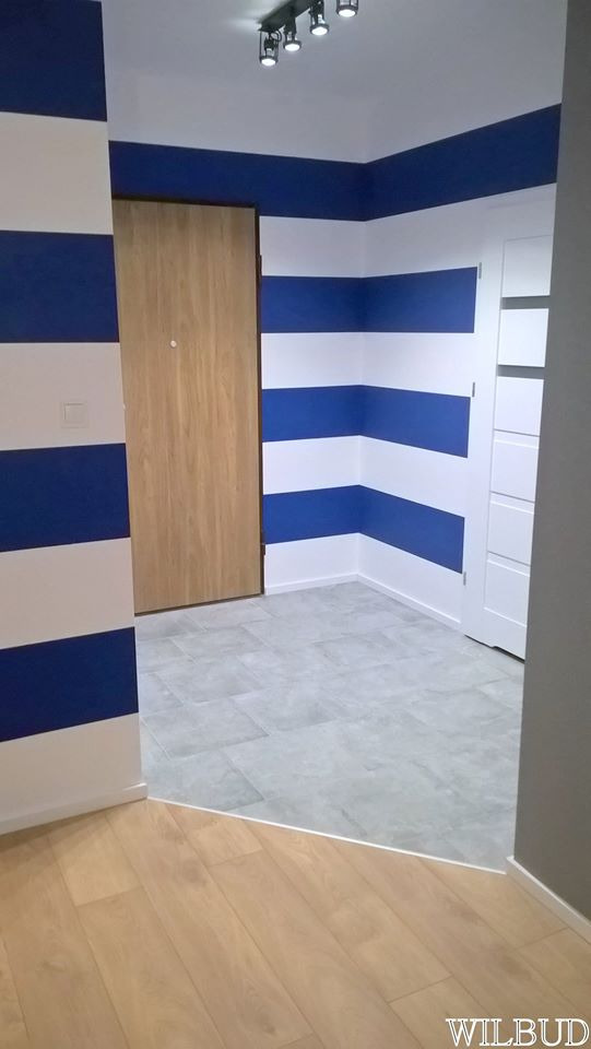 Przedpokój po remoncie w stylu marynistycznym: ściany w biało-niebieskie pasy, drewniane drzwi z wizjerem, szara podłoga z płytek i drewniana podłoga przechodząca w płytki, oświetlenie sufitowe...