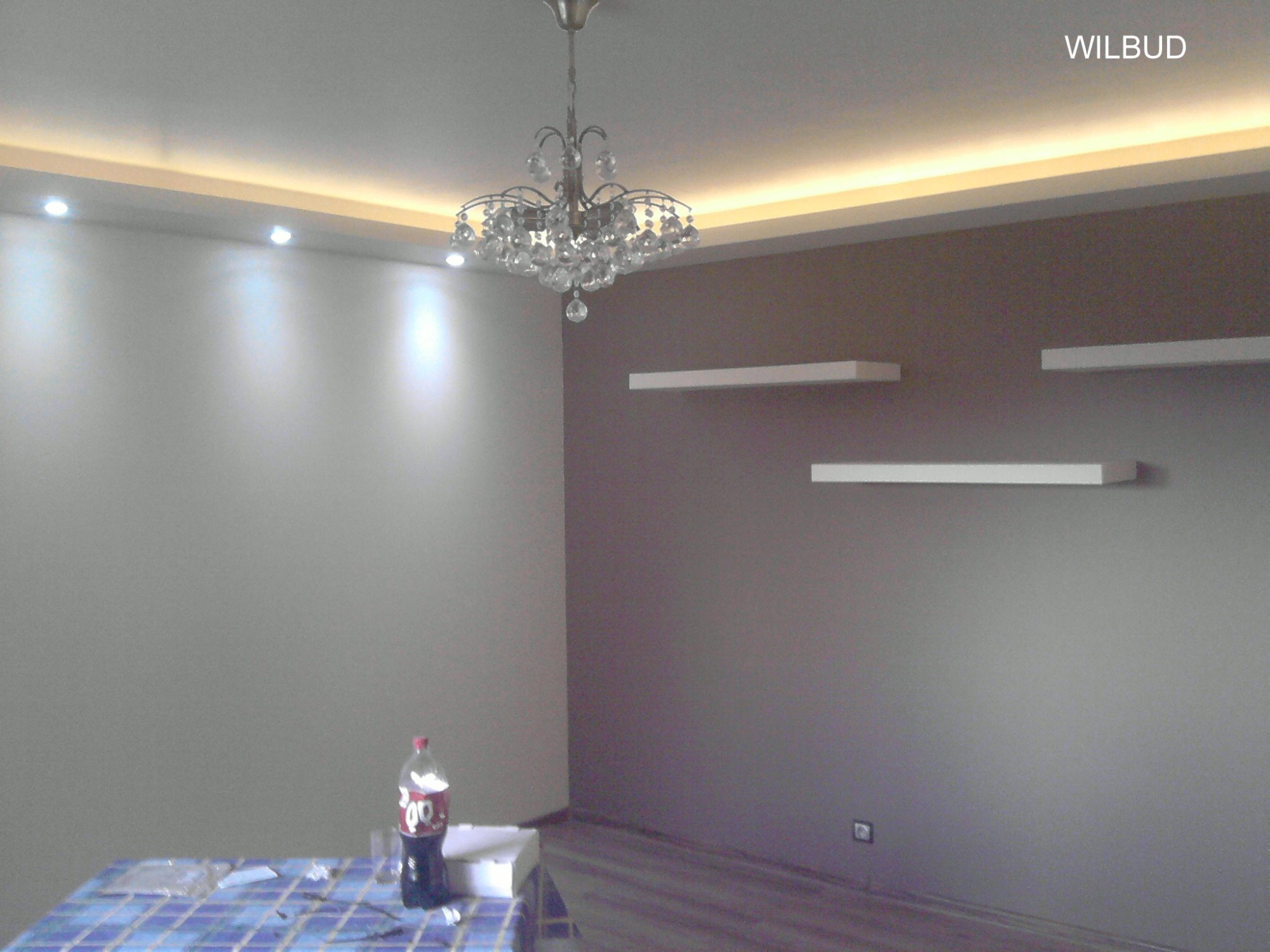 Pomalowane na dwa kolory ściany z zamontowanymi półkami i oświetleniem LED pod sufitem, widoczny żyrandol, butelka napoju na stole pod przykryciem z plamami.