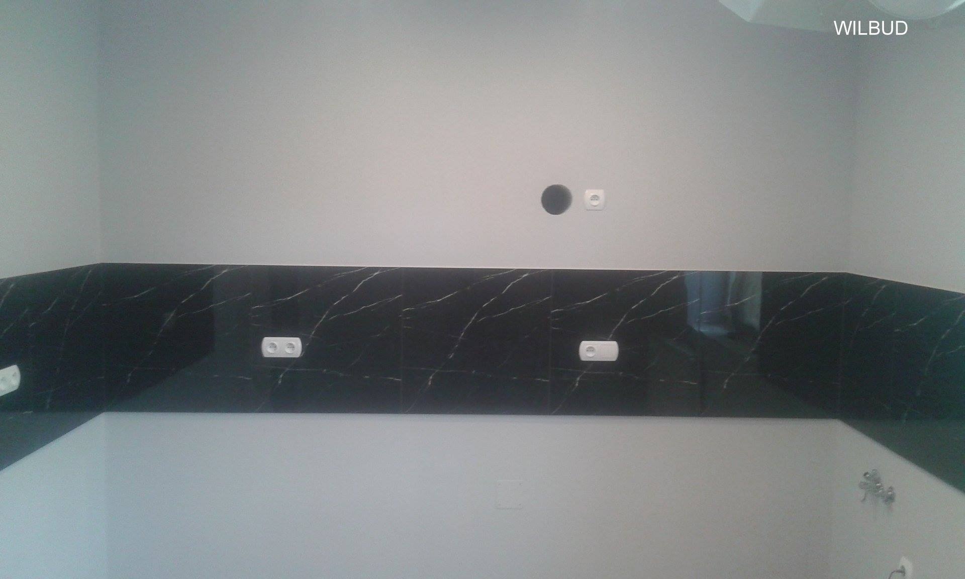 Częściowo wykończona ściana w łazience z czarnymi płytkami imitującymi marmur, widoczne gniazdka elektryczne i otwór na puszkę elektryczną, górna część ściany pomalowana na szaro.