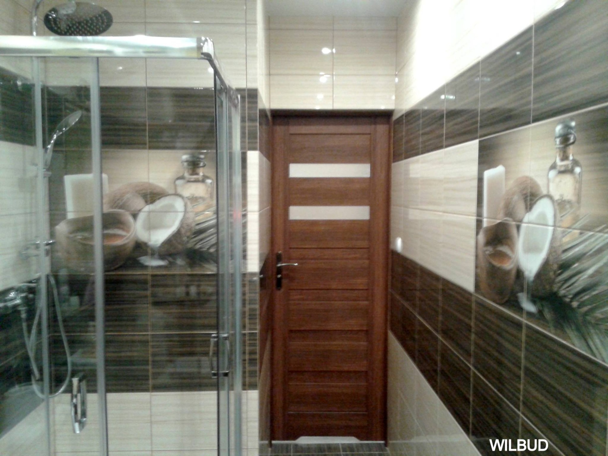 Wykończona łazienka z kabiną prysznicową i drewnianymi drzwiami. Na ścianach płytki z motywem kokosów i butelki.