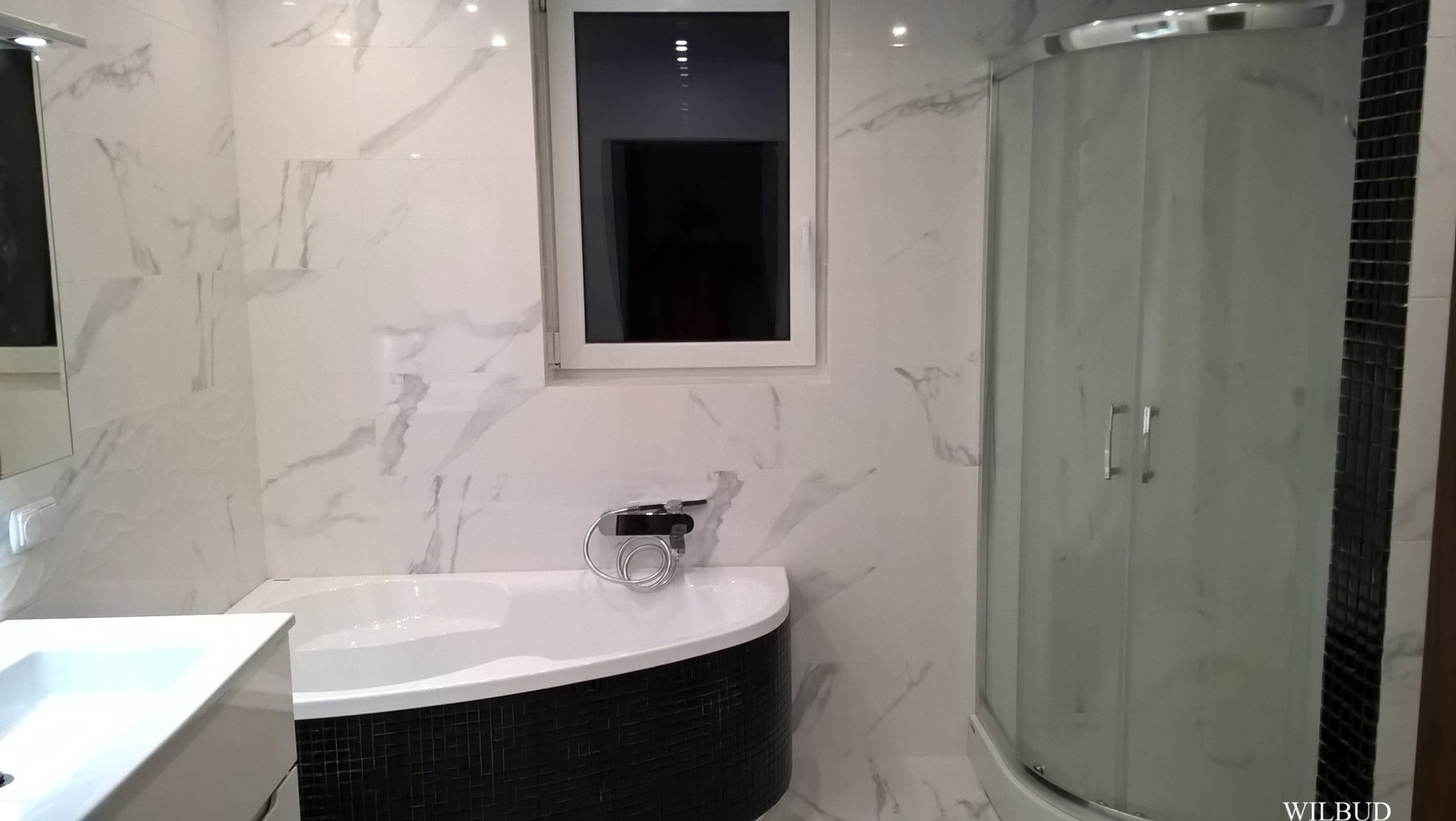 Nowoczesna łazienka z wanną obudowaną czarną mozaiką, kabiną prysznicową z matowego szkła i białymi marmurkowymi płytkami na ścianach, doświetlona przez okno.