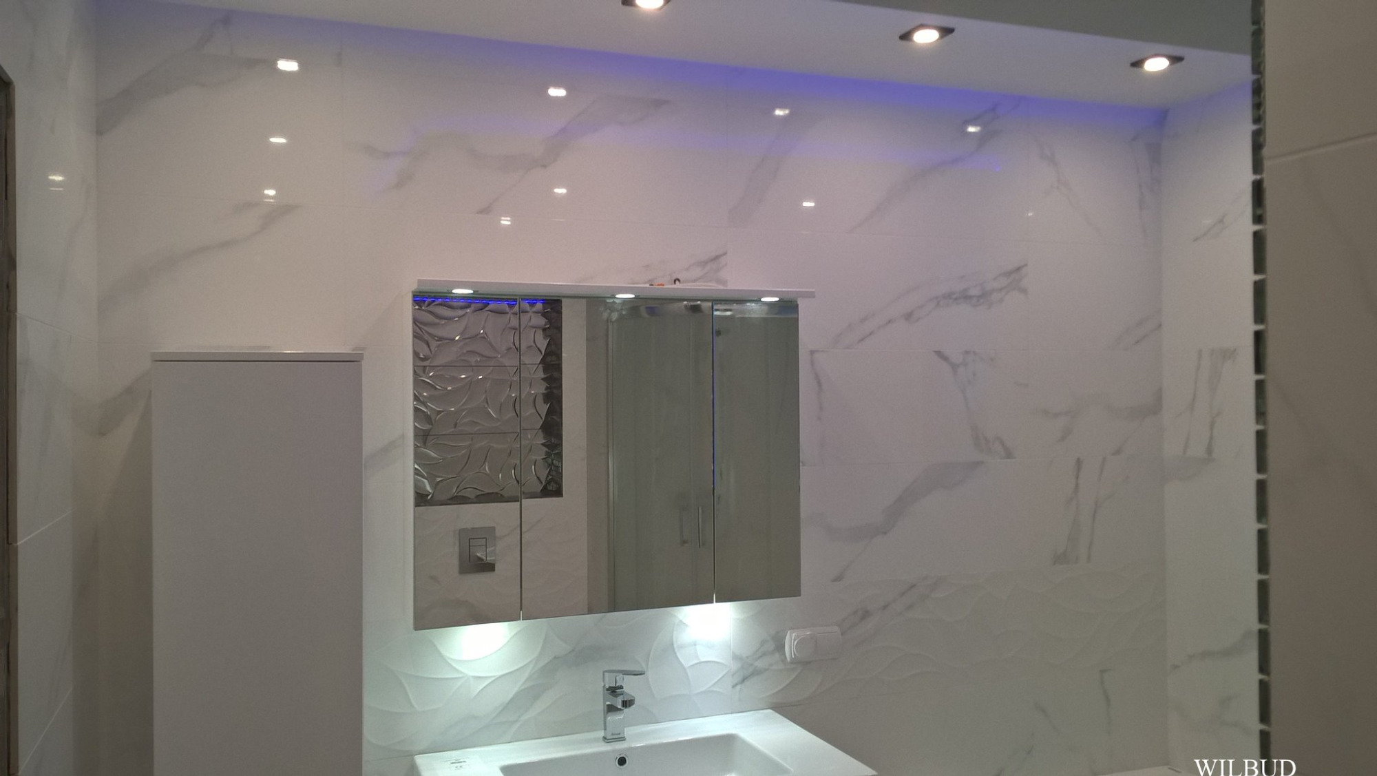 Nowoczesna łazienka z białymi płytkami imitującymi marmur, podświetlanym lustrem szafkowym, umywalką i toaletą w zabudowie.