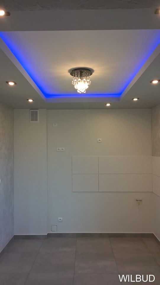 Pokój po remoncie z podwieszanym sufitem z niebieskim oświetleniem LED, centralnym żyrandolem kryształowym i punktowymi lampami halogenowymi; na ścianach gładzie i dekoracyjne panele.