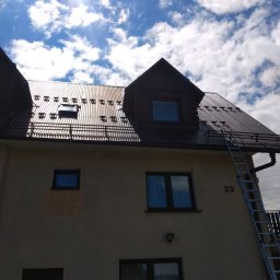 Malmal - Dom z ciemnobrązowym, metalowym dachem z licznymi odśnieżnikami, widok z dołu na tle błękitnego nieba z chmurami i jasnym słońcem, drabina oparta o ścianę budynku.