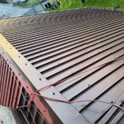 Nowy, brązowy dach z blachy trapezowej, widok z góry z fragmentem elewacji i liną zabezpieczającą