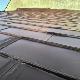 Metalowy dach pokryty świeżą, brązową farbą, widoczne łączenia blach i fragment ściany budynku w tle.