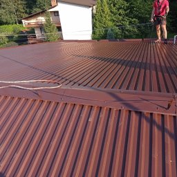 Malmal - Falista, brązowa blacha na dachu domu, na której stoi pracownik w czerwonej koszulce z uprzężą, obok wiadro farby i lina zabezpieczająca.