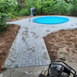 Układanie ścieżki z szarej kostki brukowej prowadzącej do okrągłego, niebieskiego basenu ogrodowego; w tle osoba pracuje w ogrodzie, a na pierwszym planie zagęszczarka do kostki.