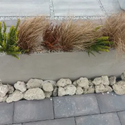 Betonowa donica z ozdobnymi trawami i krzewami, otoczona porowatymi kamieniami na szarym chodniku z kostki brukowej.