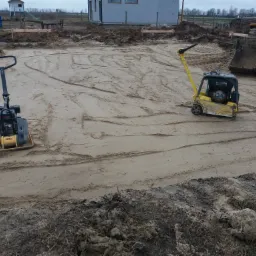 Dwa zagęszczarki gruntu, żółta i czarna, stoją na wyrównanym, piaszczystym terenie budowy, z widocznymi śladami po przejeździe maszyn, w tle budynek w trakcie budowy i fragment koparki.
