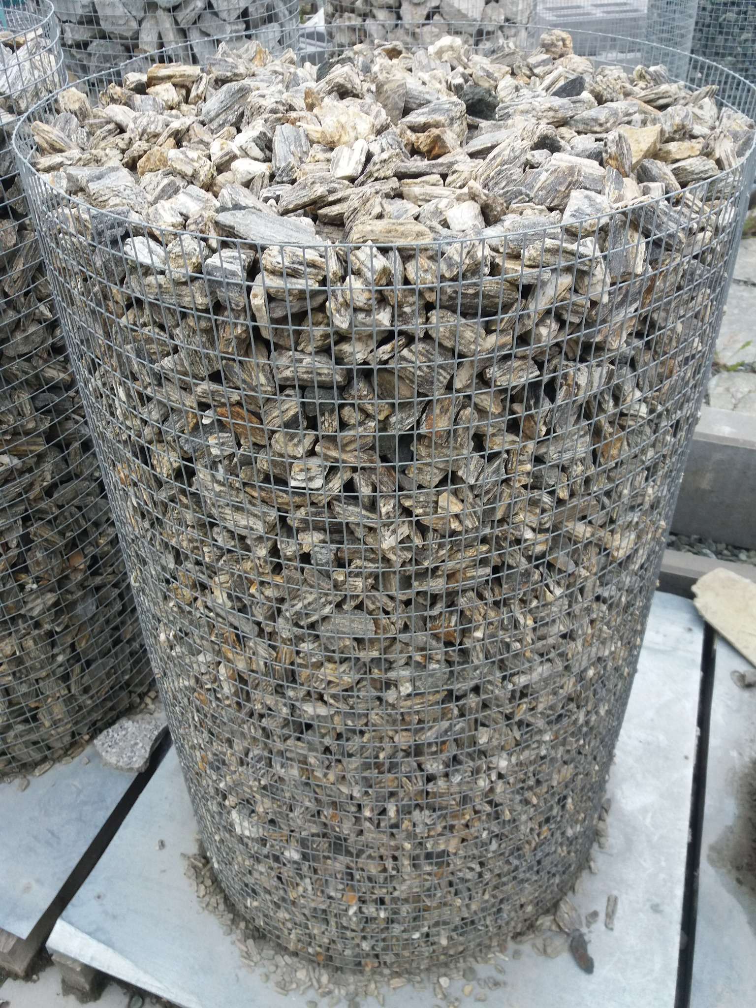 Cylindryczny kosz gabionowy wypełniony dekoracyjnym kamieniem łupanym w odcieniach szarości i beżu, widoczny na betonowej nawierzchni. Metalowa siatka kosza tworzy regularny wzór.