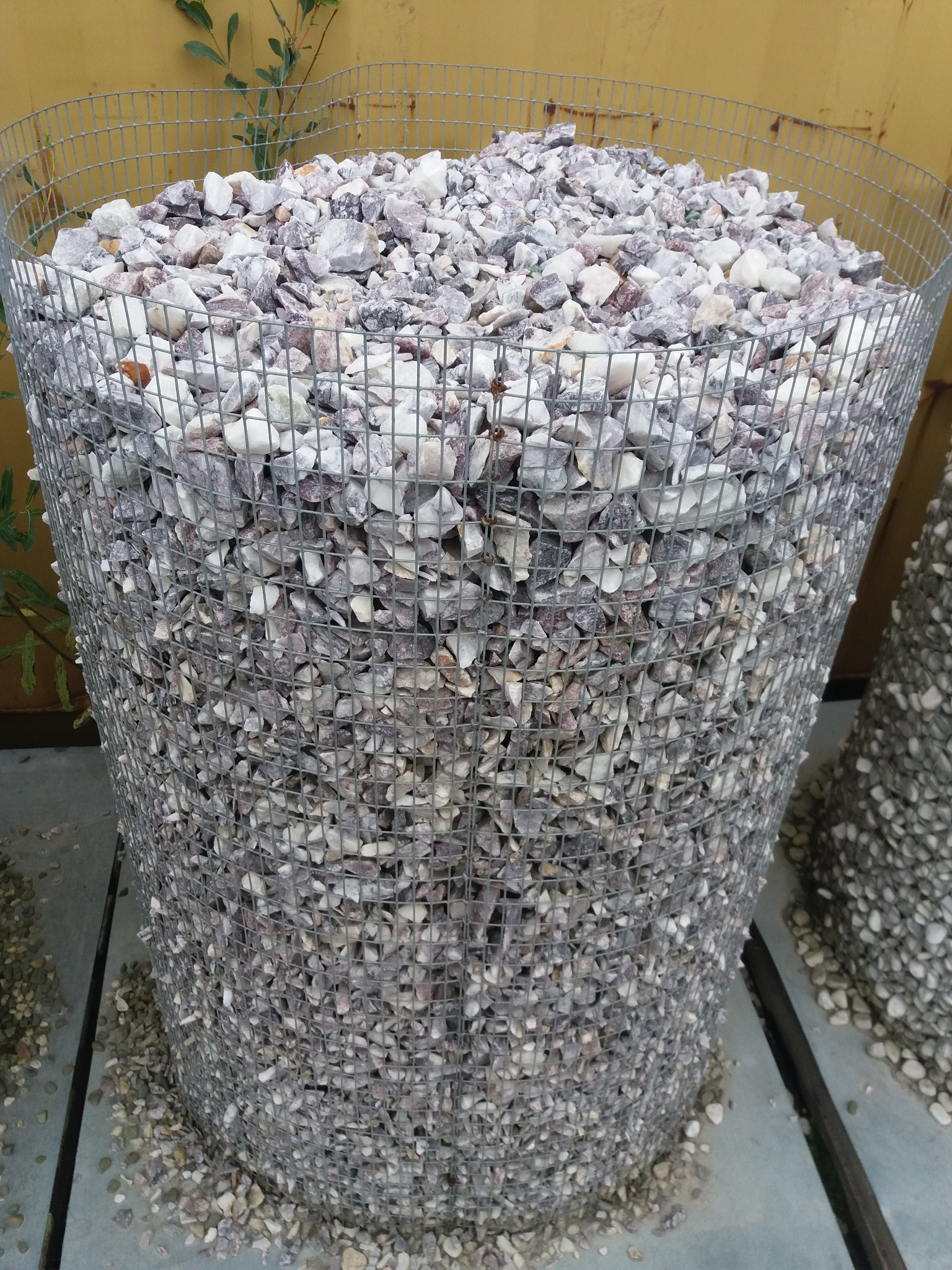Metalowa siatka wypełniona ozdobnymi kamieniami w odcieniach bieli i szarości z fioletowymi akcentami, widoczne pojedyncze kamienie na zewnątrz siatki, żółta ściana w tle.