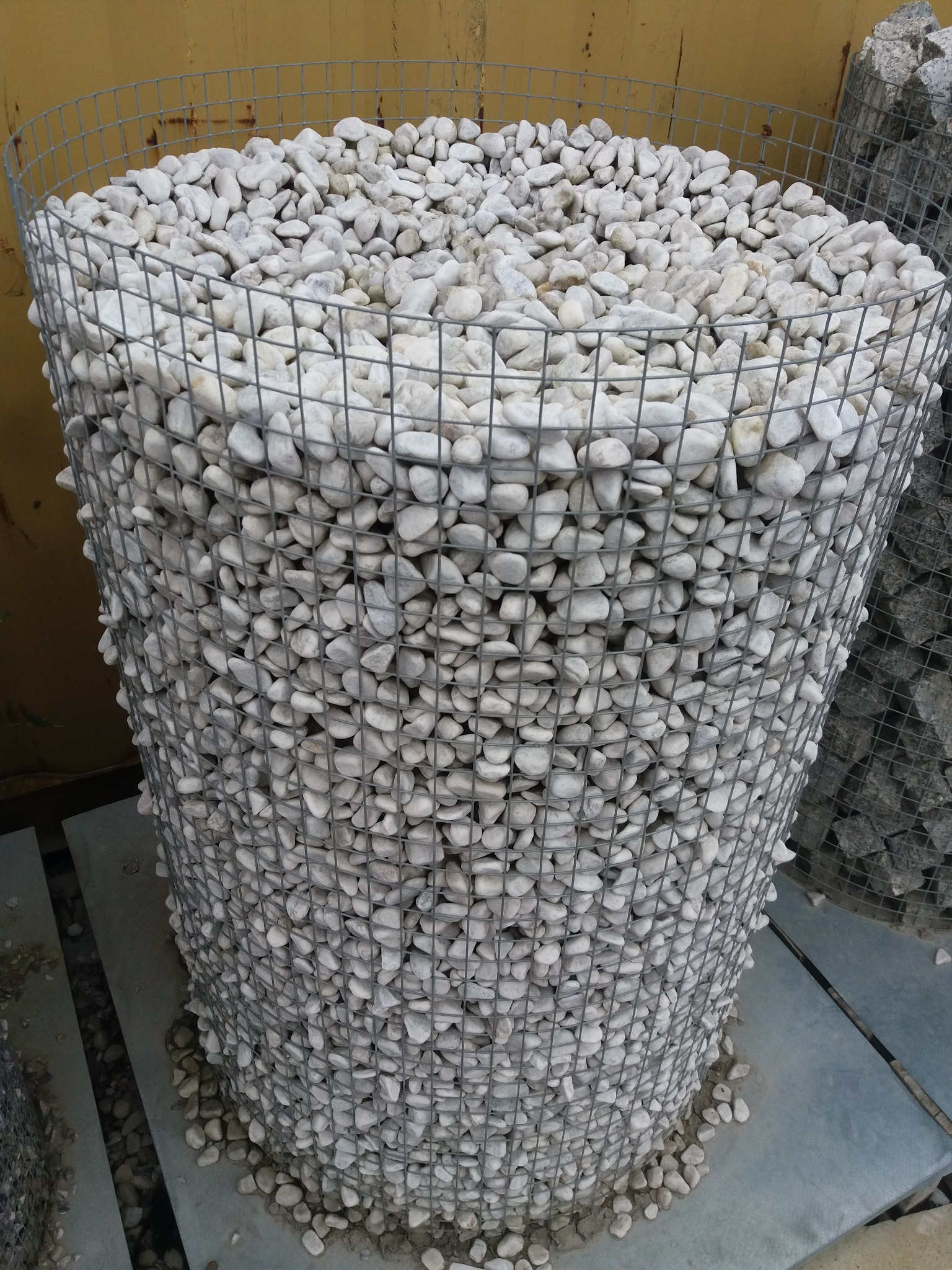 Gabion z białymi otoczakami, wypełniony po brzegi, widoczny fragment drugiego gabionu w tle, ułożony na szarych płytach.