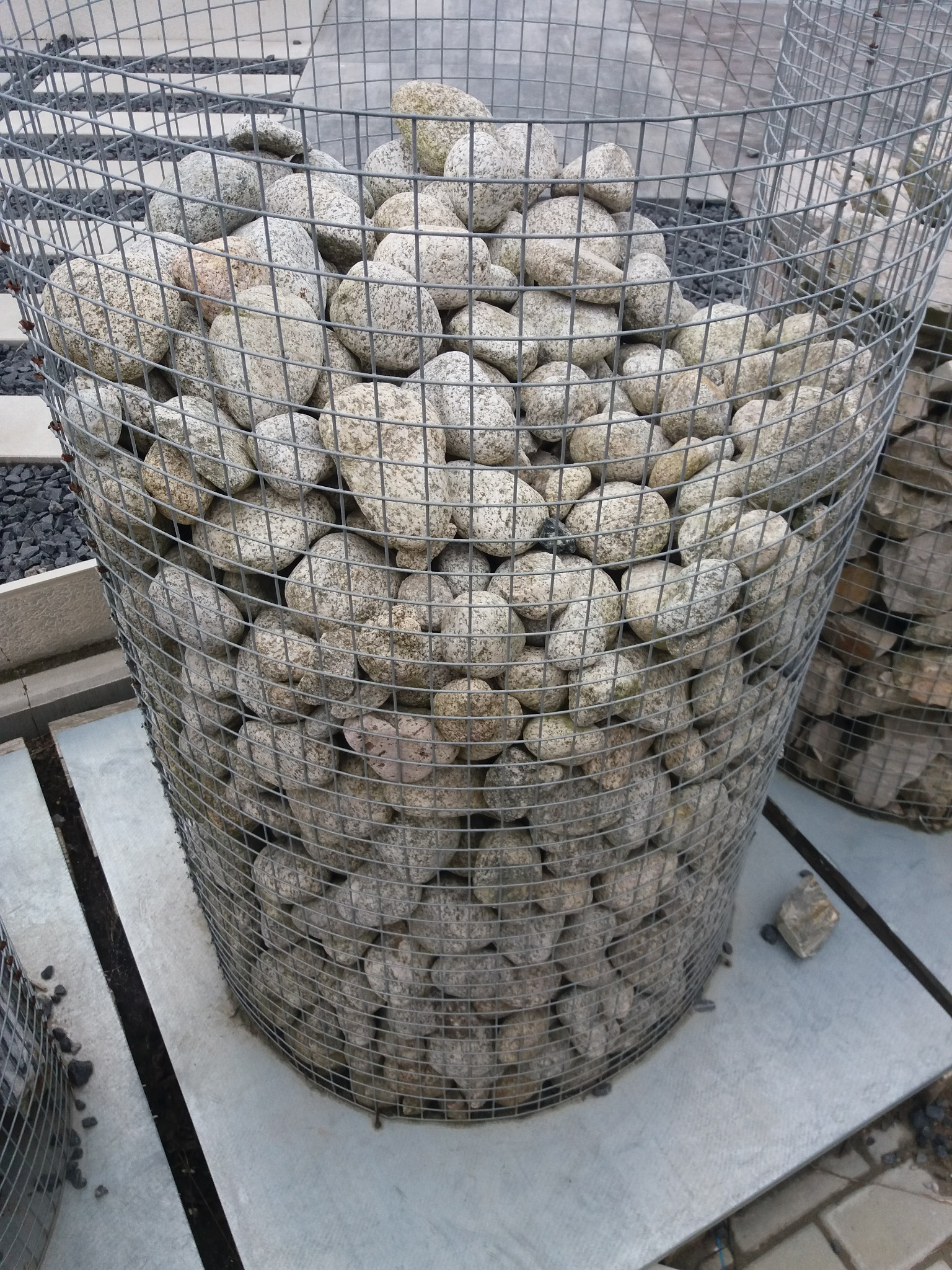 Cylindryczny gabion z szarej siatki wypełniony jasnymi, okrągłymi kamieniami, stojący na szarych płytach chodnikowych, z fragmentem drugiego gabionu w tle.