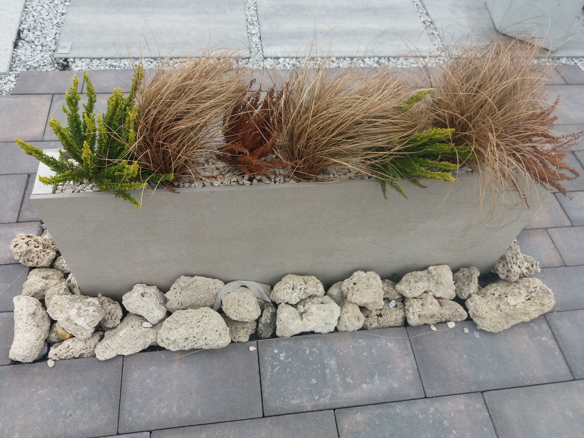 Betonowa donica z ozdobnymi trawami i krzewami, otoczona porowatymi kamieniami na szarym chodniku z kostki brukowej.