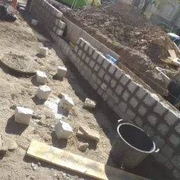 Wznoszenie niskiego muru z kostki granitowej w trakcie budowy, widoczne luźne kostki, wiadro i nasyp ziemi w tle.