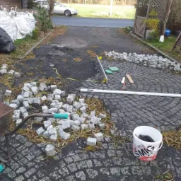 Układanie granitowej kostki w okrągły wzór na podjeździe, widoczne narzędzia, taczka z kostką i worki z materiałem budowlanym w tle.