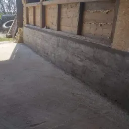 Świeżo wymurowana, surowa ściana z betonu komórkowego przy drewnianej konstrukcji budynku gospodarczego, widoczne ślady zaprawy i nierówności powierzchni, na pierwszym planie betonowa posadzka.