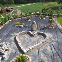 Firma prywatna - Formowanie rabaty w kształcie serca z kostki granitowej na czarnej geowłókninie, z widocznymi kamieniami, roślinami i niedokończonymi elementami ogrodu w tle.