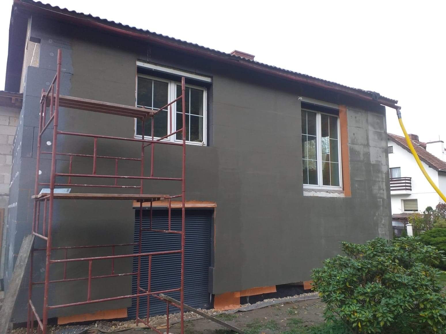 Ściana budynku ocieplana styropianem i pokrywana szarym tynkiem, widoczne rusztowanie, okna z białymi ramami i częściowo otynkowana ściana sąsiedniego budynku.