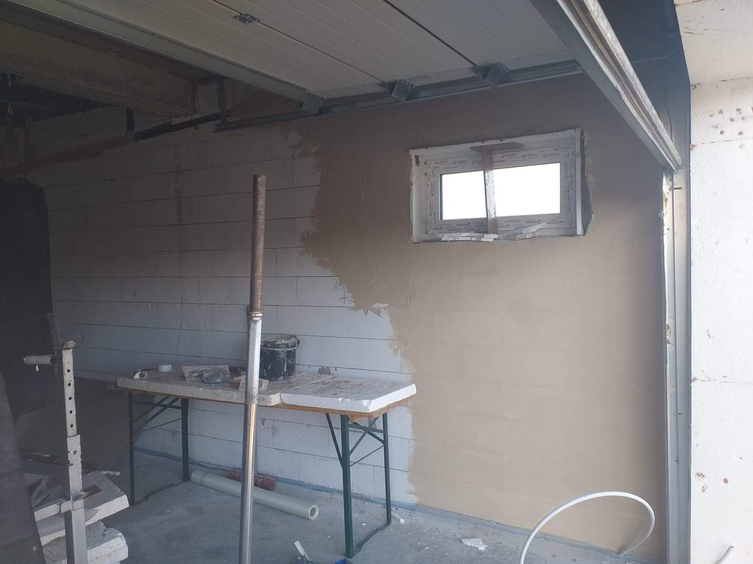 Wnętrze garażu z częściowo otynkowaną ścianą z bloczków, widoczne okno z folią ochronną, stół roboczy z narzędziami i wiadro, wspornik sufitowy bramy garażowej.