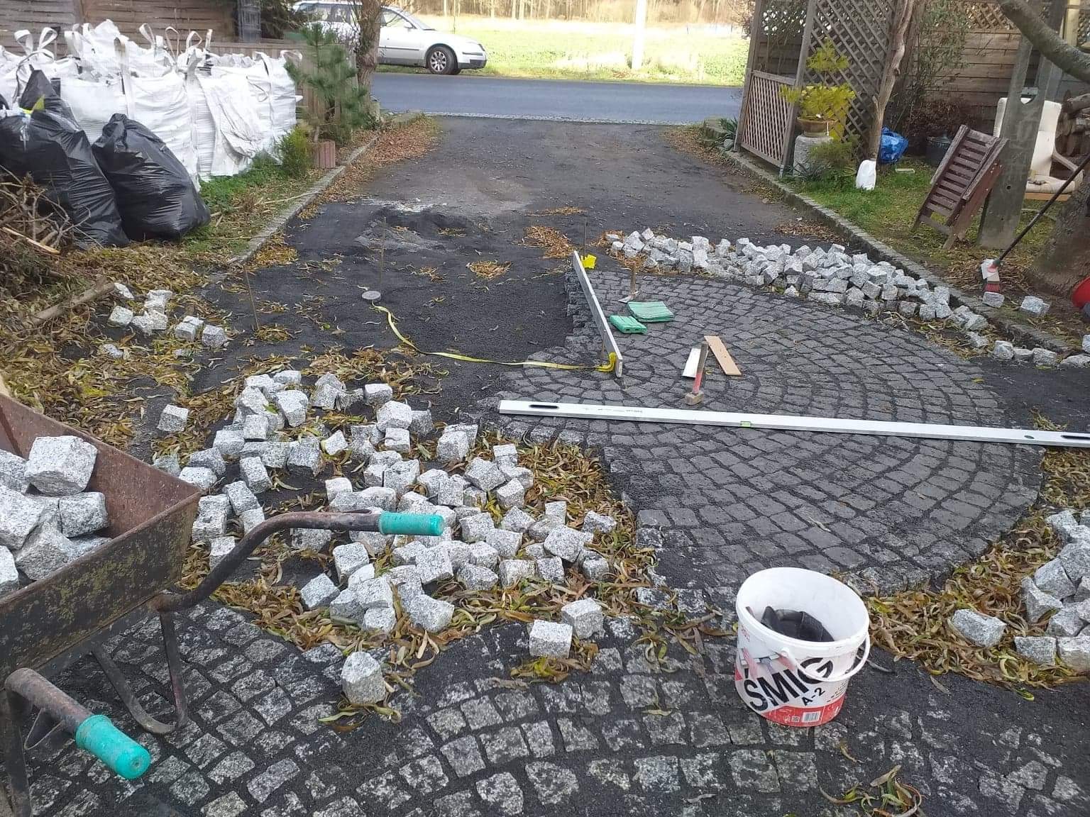 Układanie granitowej kostki w okrągły wzór na podjeździe, widoczne narzędzia, taczka z kostką i worki z materiałem budowlanym w tle.