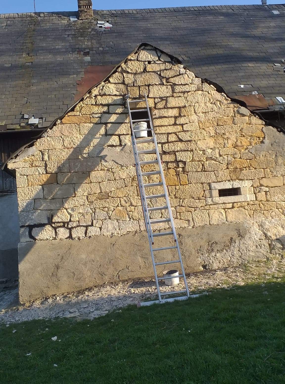 Kamienna ściana budynku w trakcie renowacji, z widocznymi ubytkami i nowym tynkiem u podstawy, oparta o nią drabina, dwa plastikowe wiadra na różnych poziomach, w tle uszkodzony dach z dachówki.