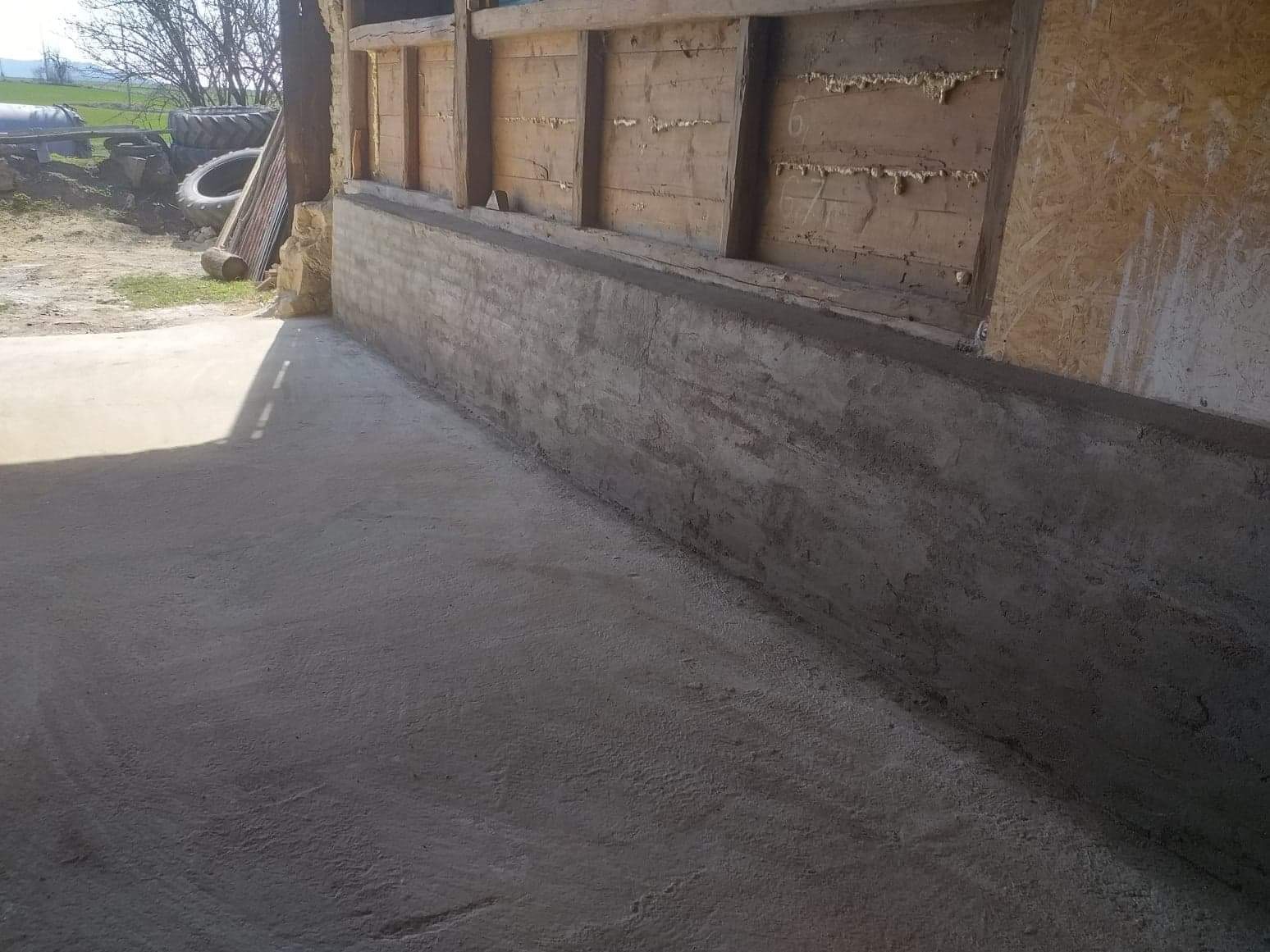 Świeżo wymurowana, surowa ściana z betonu komórkowego przy drewnianej konstrukcji budynku gospodarczego, widoczne ślady zaprawy i nierówności powierzchni, na pierwszym planie betonowa posadzka.