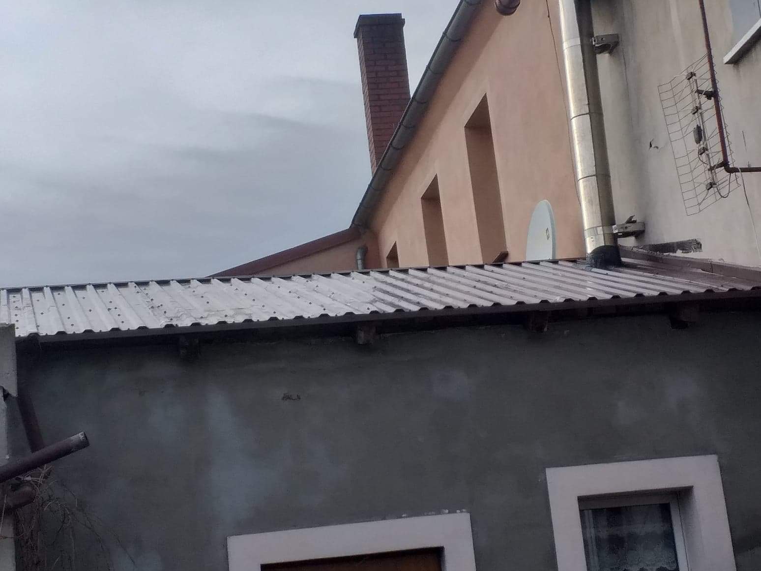 Szara ściana budynku z dwoma oknami w białych ramach, nad nią blaszany dach z widocznymi śladami korozji, w tle komin z czerwonej cegły, antena satelitarna i przewody kominowe ze stali nierdzewnej.