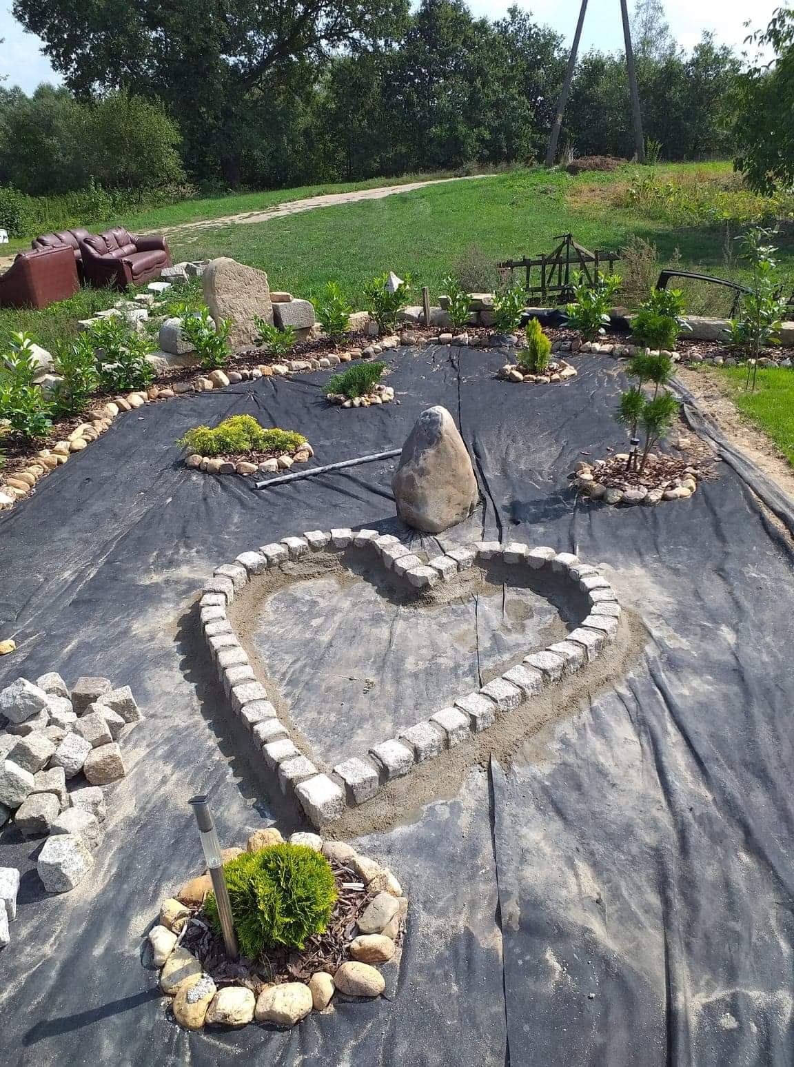 Formowanie rabaty w kształcie serca z kostki granitowej na czarnej geowłókninie, z widocznymi kamieniami, roślinami i niedokończonymi elementami ogrodu w tle.