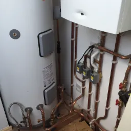 Instalacja grzewcza z białym zasobnikiem Megaflo i piecem, widoczne miedziane rury, zawory i skrzynki elektryczne na ścianie, podłoga wyłożona płytami OSB.