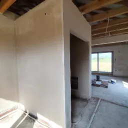 Wnętrze budynku w trakcie wykańczania, ściany pokryte świeżo nałożonym, jasnym tynkiem, widoczne drewniane elementy konstrukcji dachu i wystające przewody elektryczne.