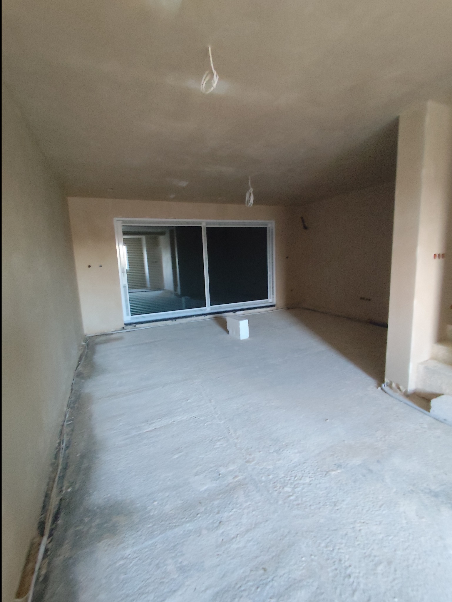 Wnętrze pomieszczenia po nałożeniu tynków, widoczne świeżo otynkowane ściany i sufit, z wystającymi kablami elektrycznymi, oraz duże okno przesuwne z widokiem na kolejne pomieszczenie.