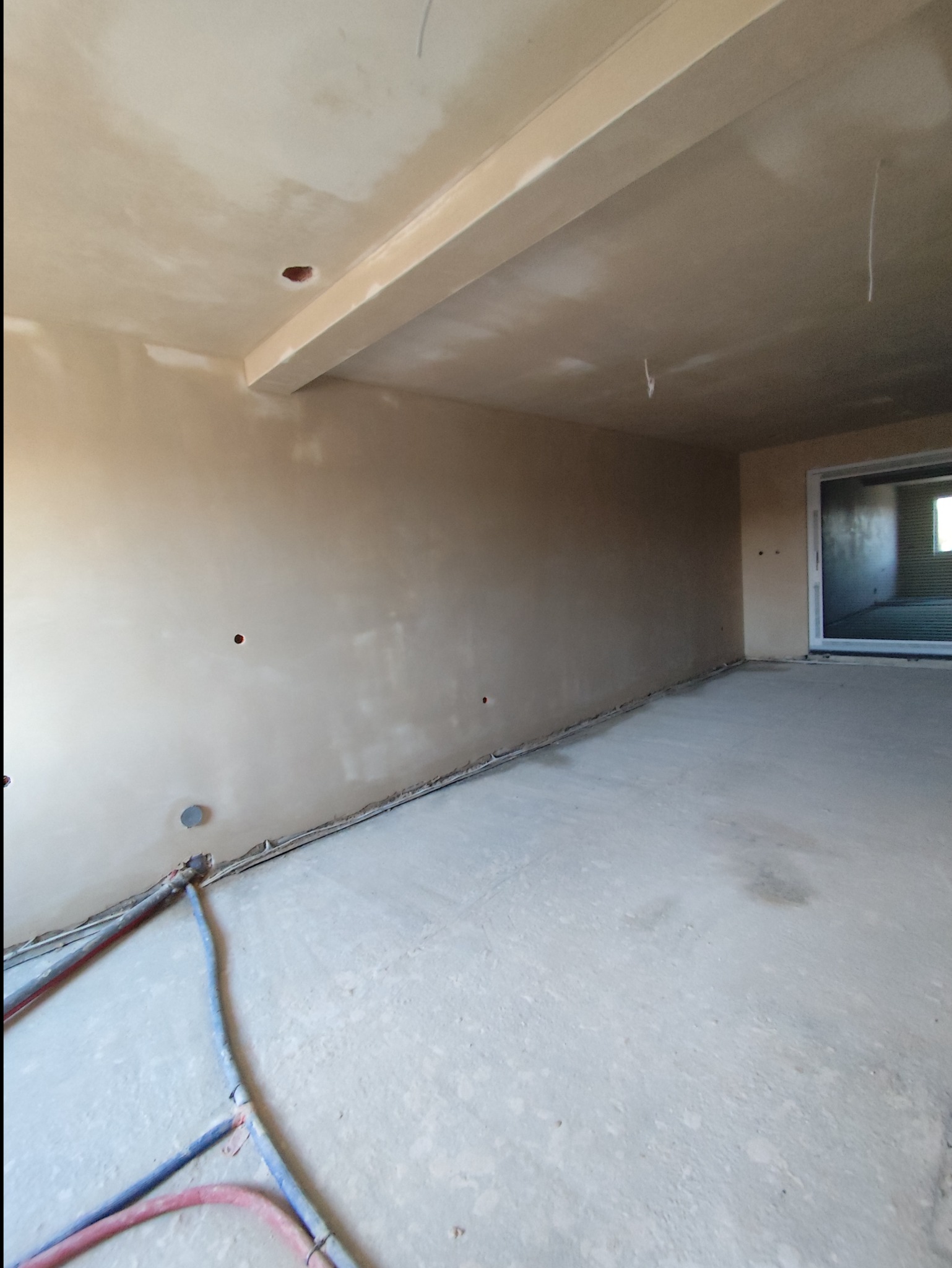 Wnętrze po nałożeniu tynków maszynowych, widoczne ściany i sufit z gładką, jasną powierzchnią, wystające rury instalacyjne przy podłodze, surowy stan wykończenia przed dalszymi pracami.