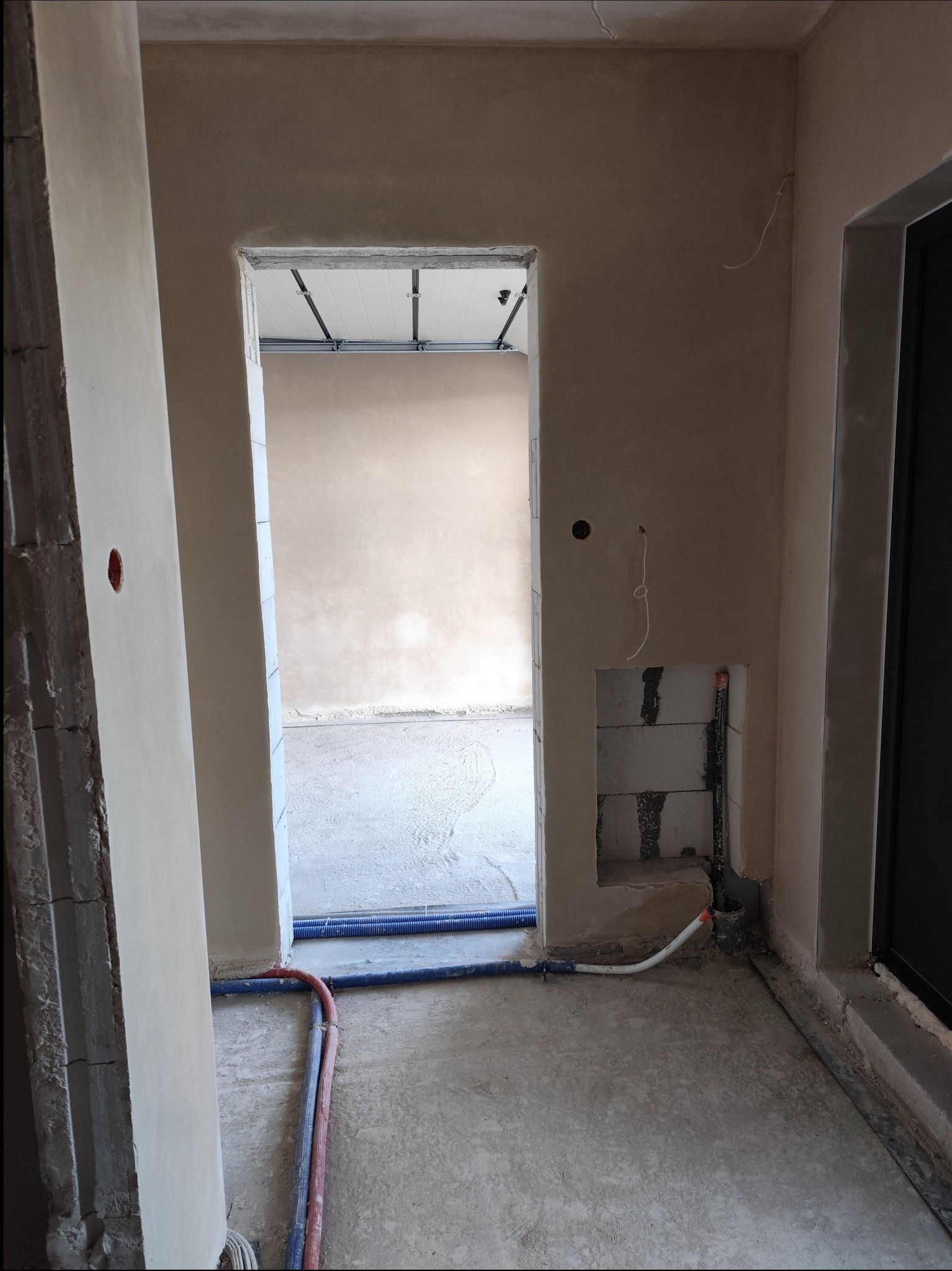 Wnętrze w trakcie prac wykończeniowych: ściany pokryte gładzią, widoczne instalacje wodne i elektryczne, surowa podłoga, otwarte przejście do kolejnego pomieszczenia z widoczną konstrukcją sufitu...