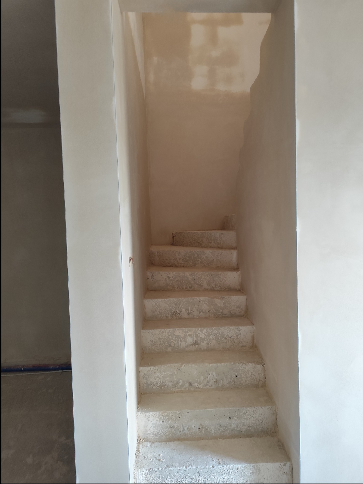 Wnętrze klatki schodowej z surowymi, betonowymi schodami po nałożeniu gładzi gipsowej na ściany, widoczne ślady po łatach na ścianach.