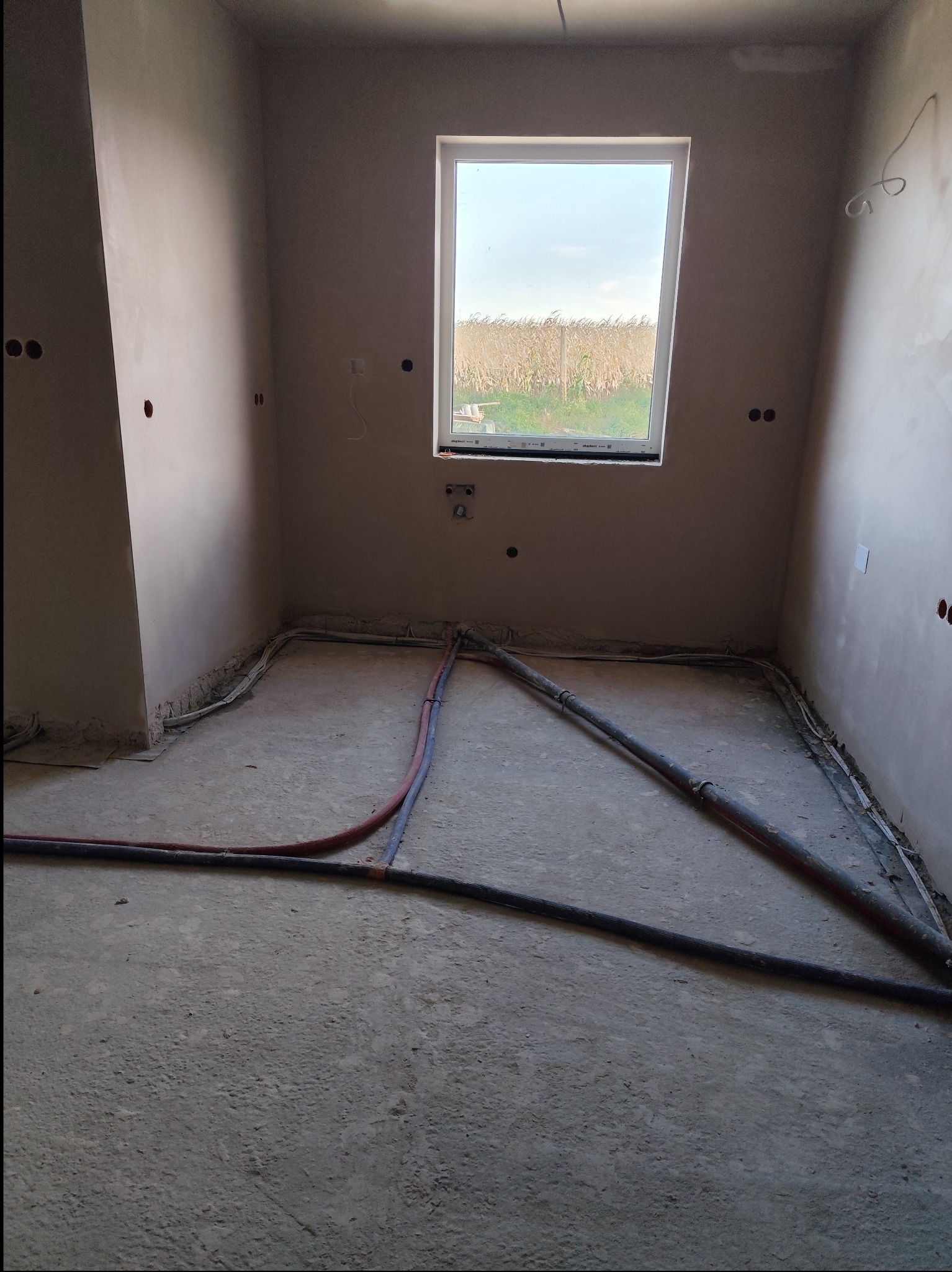Pokój po nałożeniu tynku maszynowego na ściany, widoczne przewody instalacji elektrycznej i hydraulicznej na podłodze przed wylewką, okno z widokiem na pole kukurydzy.