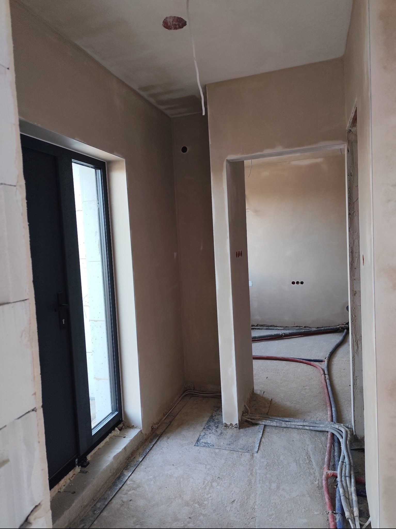 Wnętrze w trakcie prac wykończeniowych: świeżo otynkowane ściany w odcieniach beżu, widoczne instalacje elektryczne i hydrauliczne na podłodze, otwór drzwiowy z czarną ramą i widokiem na zewnątrz.