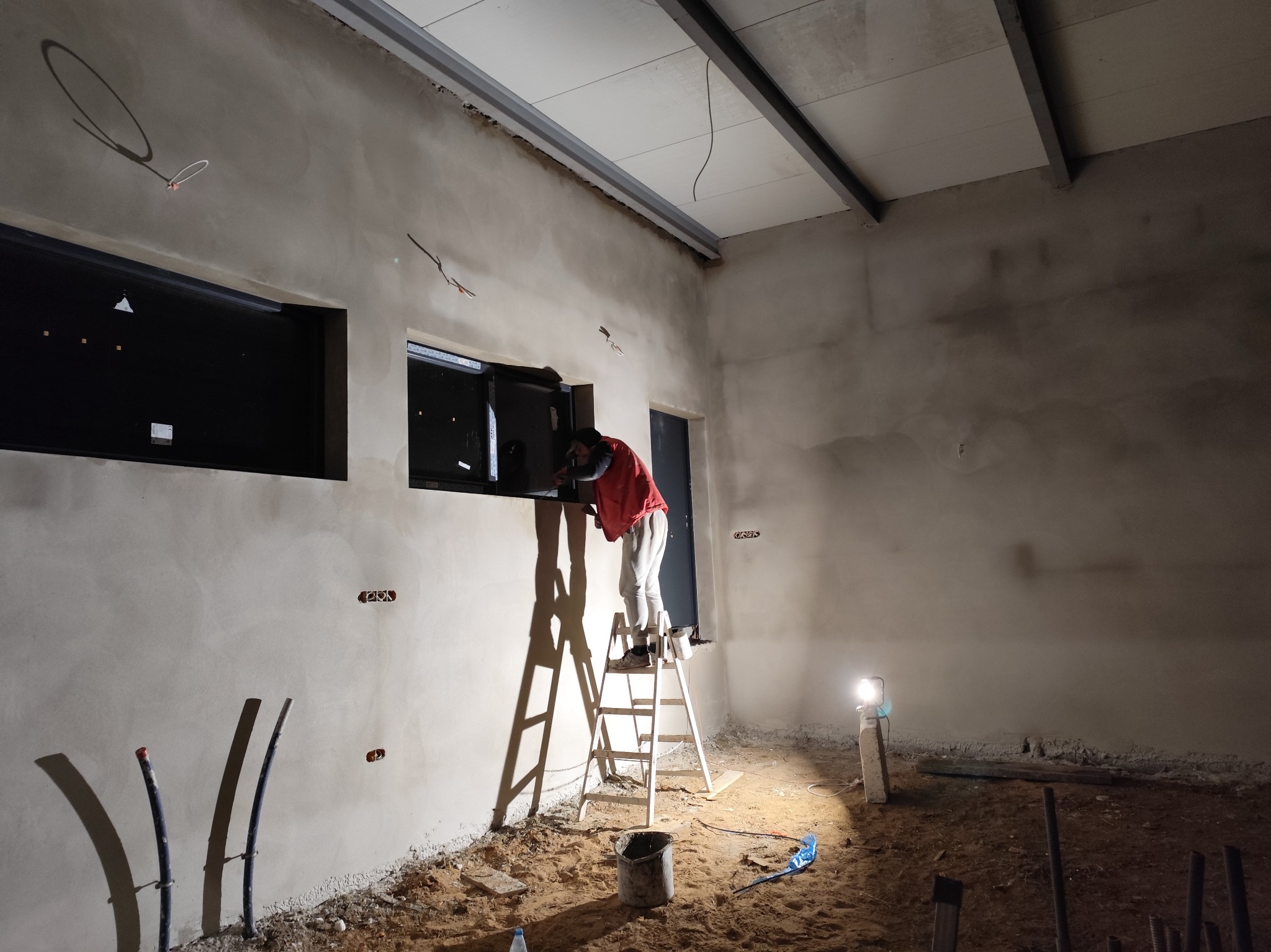 Tynkowanie maszynowe ściany wokół okna: pracownik na drabinie w czerwonej bluzie, widoczne ślady po instalacji elektrycznej, piasek na podłodze, lampa halogenowa oświetla wnętrze.
