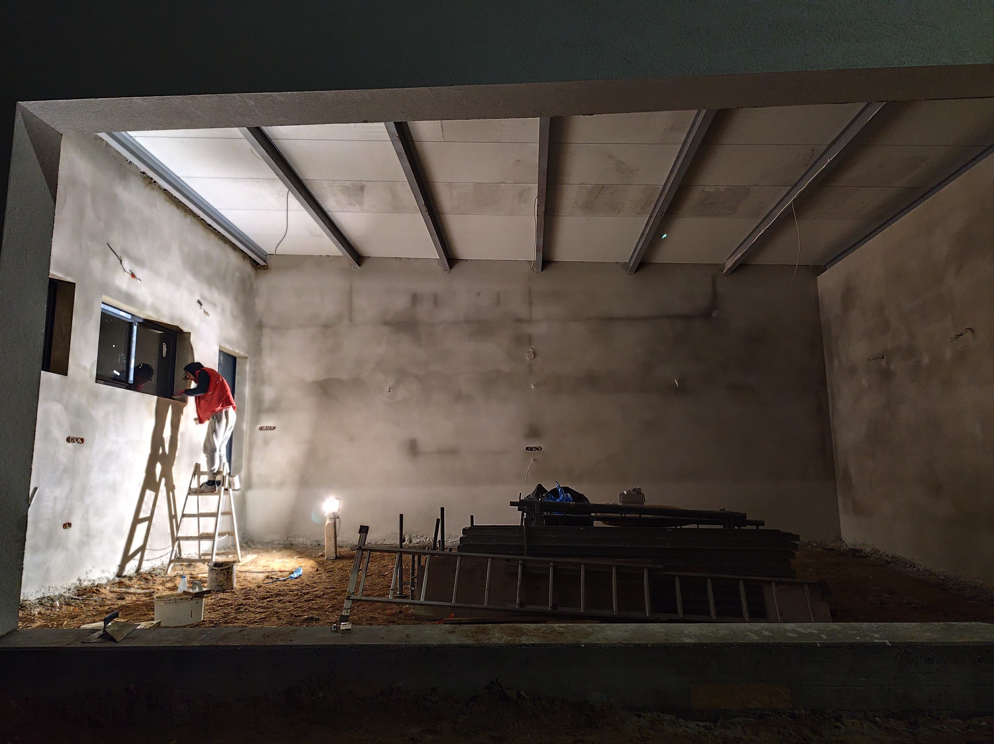 Pracownik w czerwonej kamizelce na drabinie, wygładza świeżo nałożony tynk przy oknie w surowym wnętrzu, widoczne metalowe profile sufitowe i porozrzucane narzędzia.