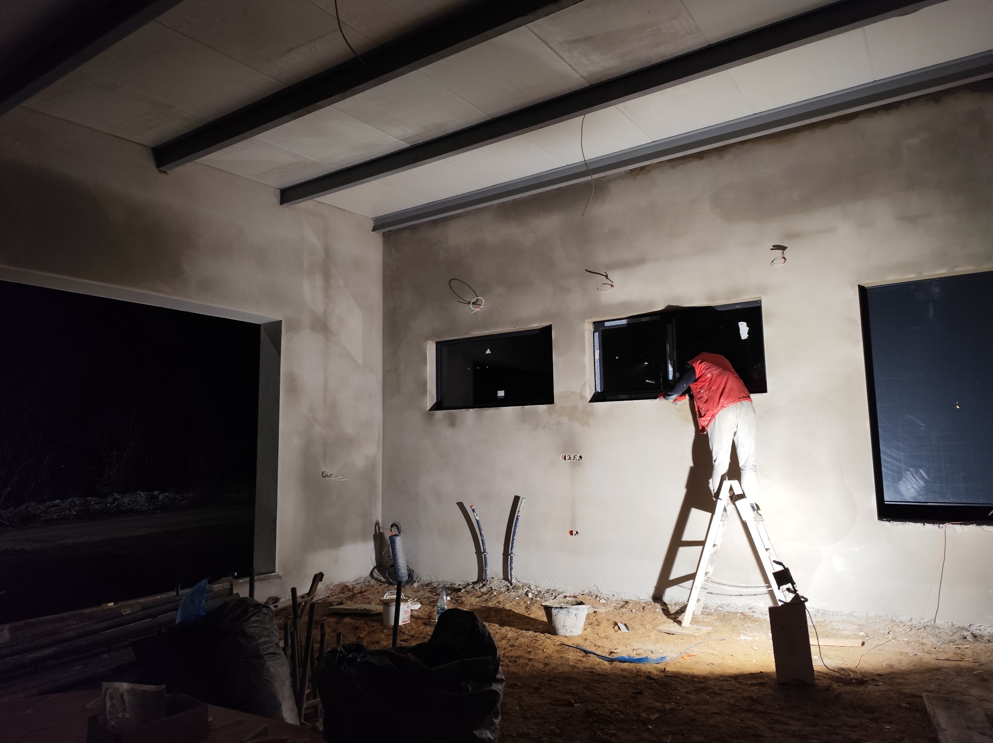 Wnętrze w trakcie prac wykończeniowych: ściany po nałożeniu gładzi, pracownik na drabinie w czerwonej kurtce, okna w czarnych ramach, widoczne instalacje elektryczne i hydrauliczne, podłoga...