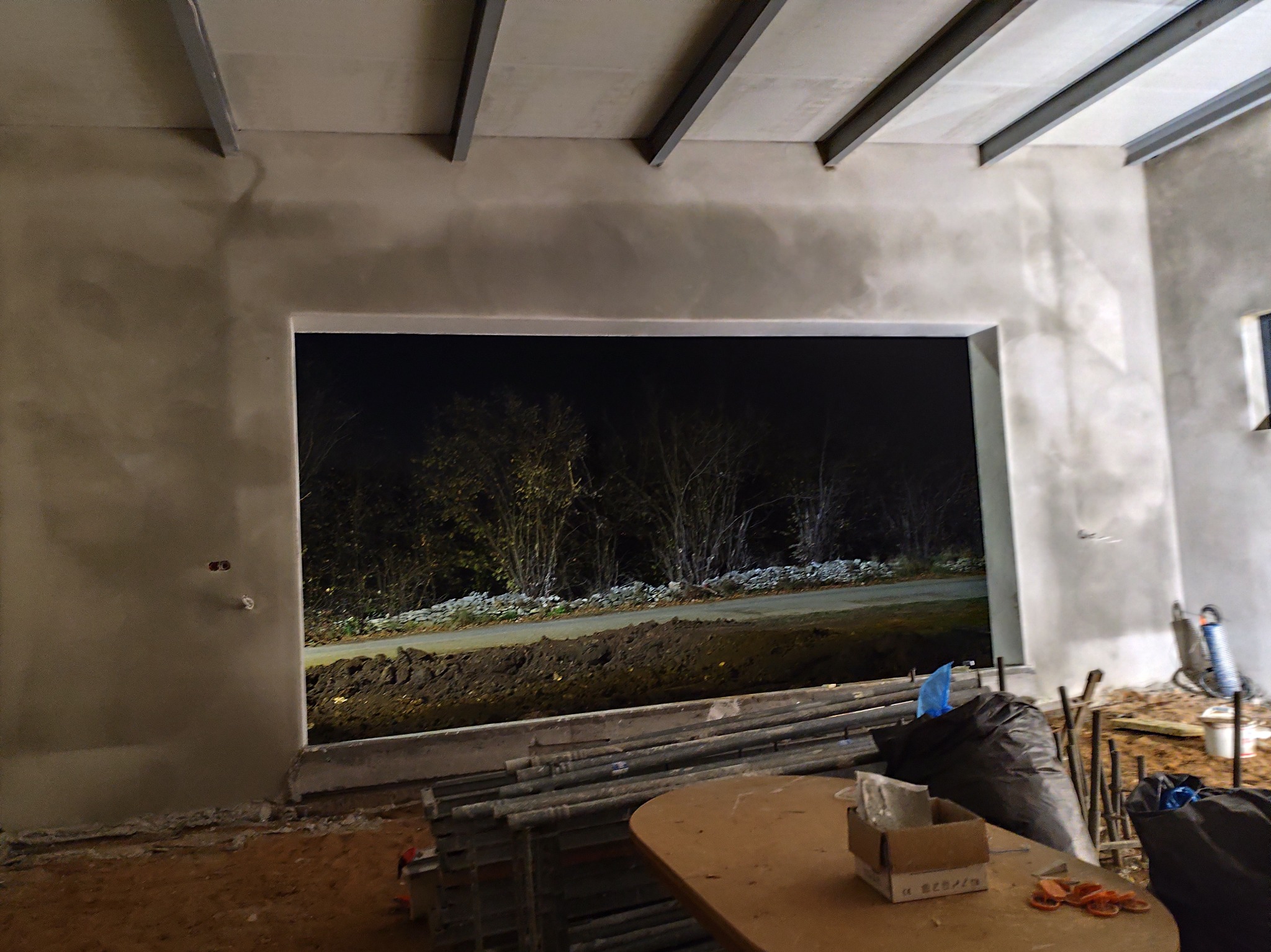 Surowy, szary tynk na ścianach wewnętrznych budynku w trakcie prac wykończeniowych, widoczne metalowe profile stropowe i widok na zewnątrz przez otwór okienny z ciemnym tłem drzew, na pierwszym...